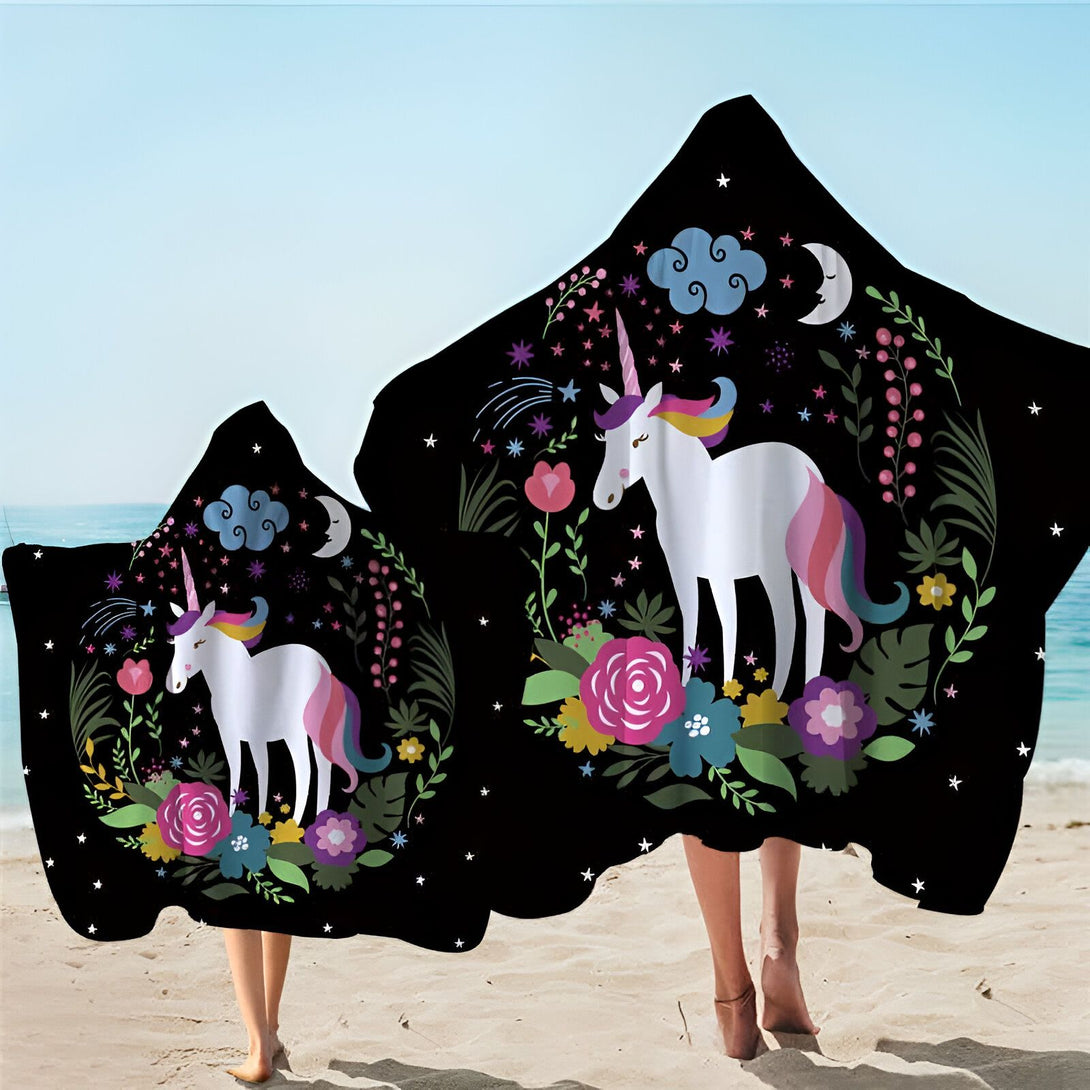 Deux personnes sur la plage portant des serviettes à capuche noires avec un motif de licorne blanche, des fleurs colorées, des étoiles, un croissant de lune et des nuages. Idéal pour les enfants et les adultes après la baignade., 45501226090751 | Licorne Shop