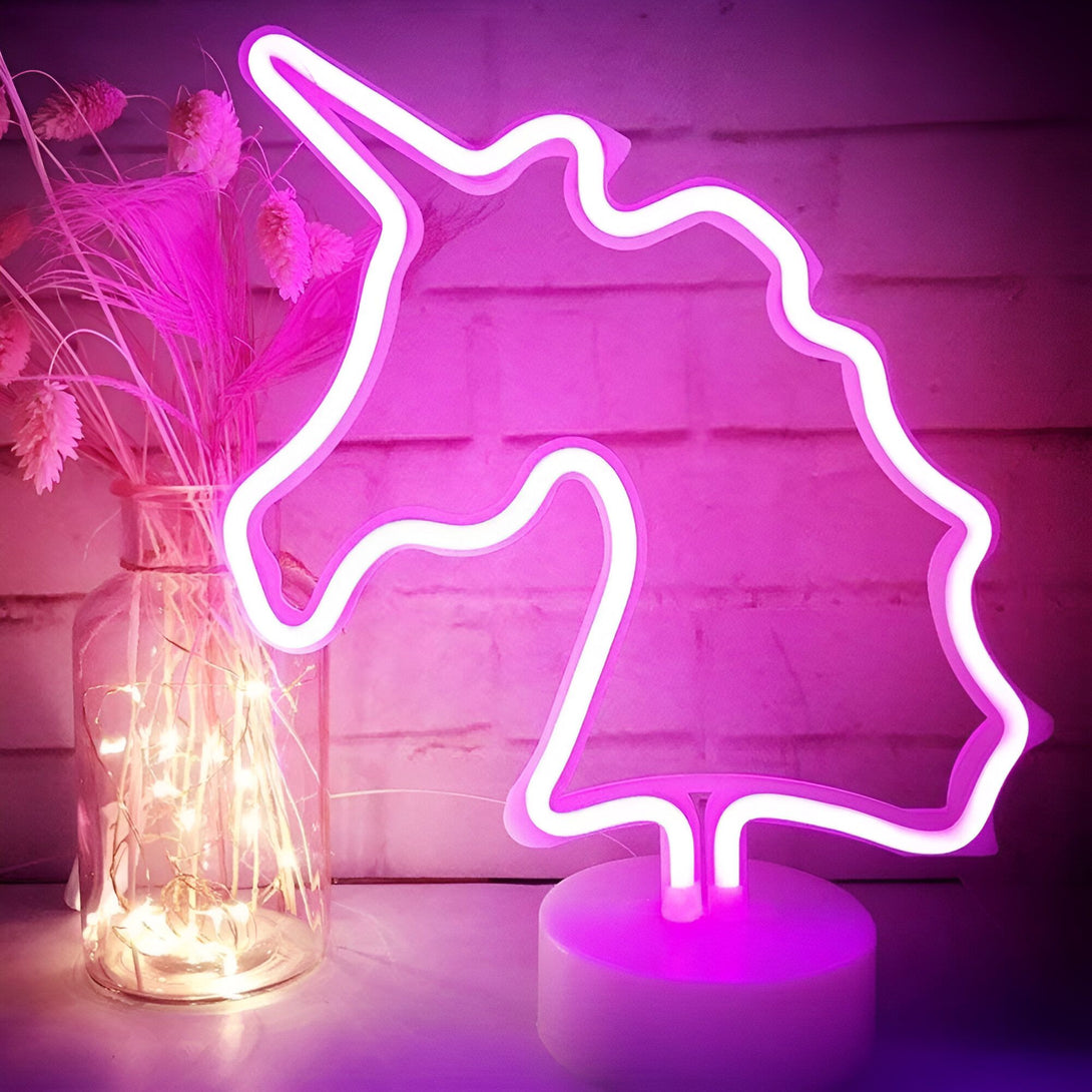 Une enseigne lumineuse en forme de licorne rose est posée sur une table. A côté d'elle, se trouve un vase en verre transparent contenant des fleurs séchées et une guirlande lumineuse. Le tout est posé sur un mur en briques blanches.