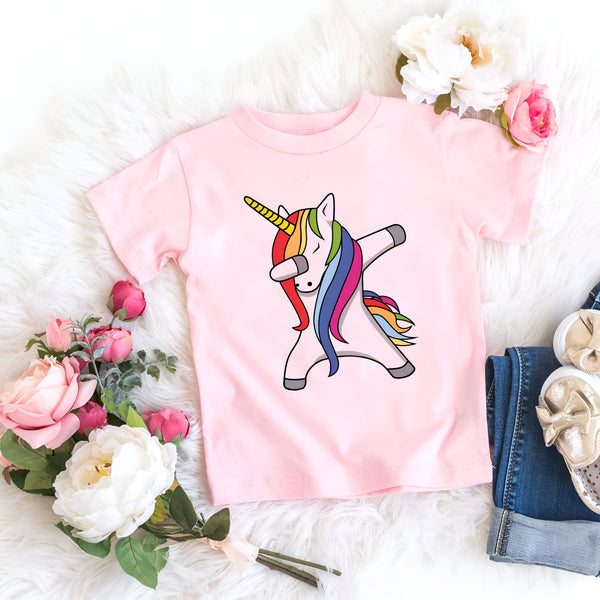 T-shirt rose pour enfant avec un motif de licorne arc-en-ciel faisant un "dab". Des fleurs roses et blanches, un jean retroussé et des chaussures dorées complètent la scène sur une couverture en fausse fourrure blanche., 45502325227775,45502325260543,45502325293311,45502325326079,45502325358847,45502325391615,45502325424383,45502325457151 | Licorne Shop