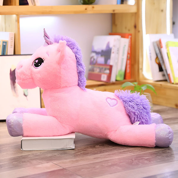 Peluche licorne rose et violette allongée sur un livre. Crinière et queue violettes, petit cœur brodé sur le flanc. Étagère en bois avec des livres en arrière-plan. Ambiance douce et enfantine., 45501127819519 | Licorne Shop