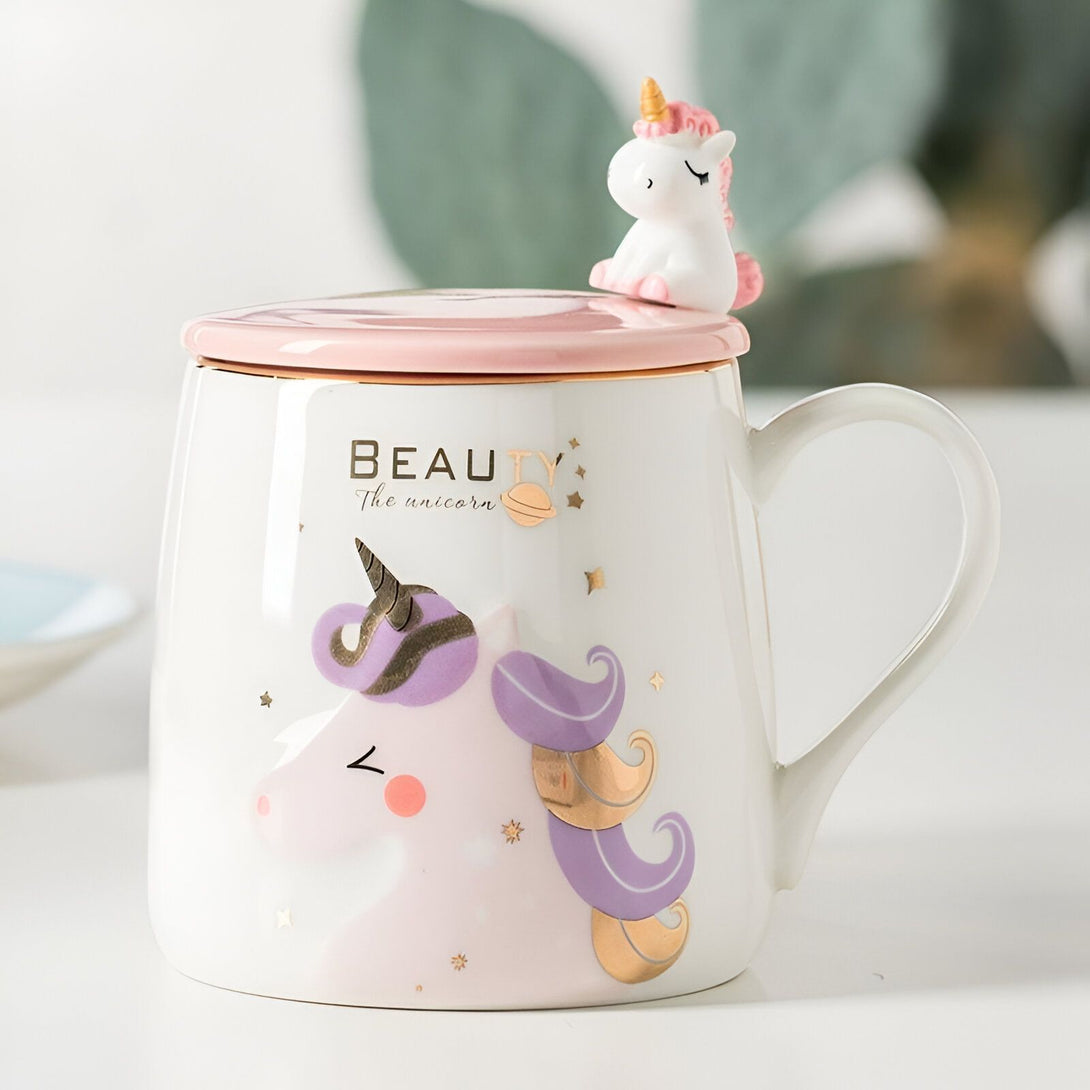 Une tasse en céramique blanche avec un couvercle rose et une cuillère en forme de licorne. La tasse est décorée d'une illustration d'une licorne violette et dorée avec des étoiles et des lunes.