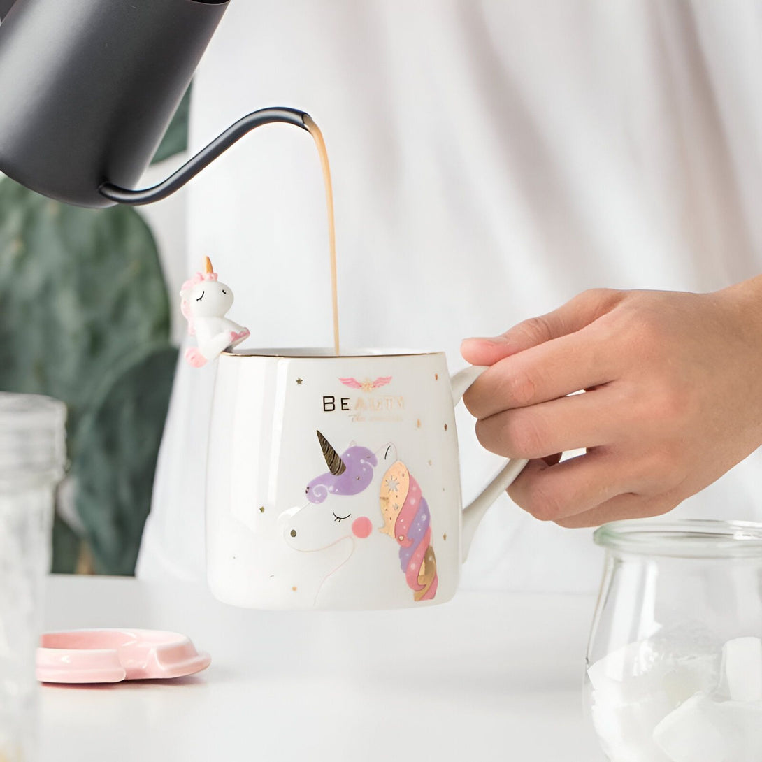 Une tasse en céramique blanche avec un motif de licorne. La tasse a une anse et un couvercle. Il y a une petite figurine de licorne sur le bord de la tasse. La tasse est remplie de thé.