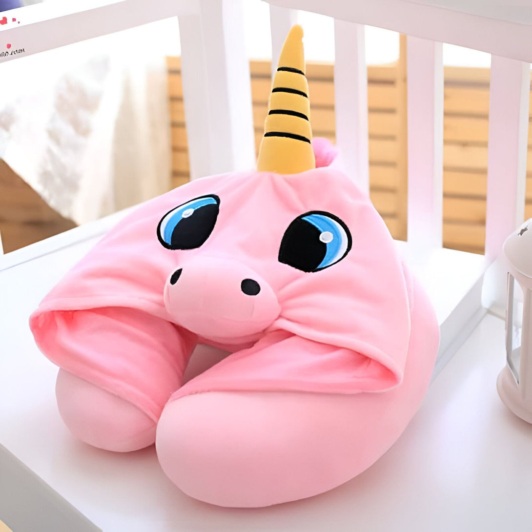 Image d'un coussin de voyage en forme de licorne rose avec des yeux bleus, un nez noir et une corne jaune à rayures noires. Le coussin repose sur une surface blanche devant un fond de barrières blanches et un objet blanc sur le côté droit., 45501060841727 | Licorne Shop