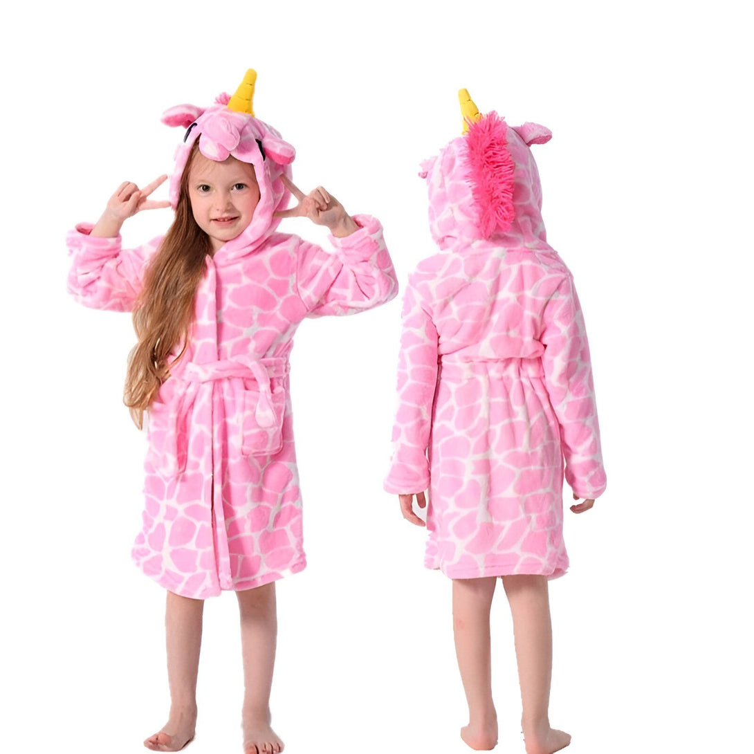 Une jeune fille porte une combinaison pyjama rose à capuche en forme de licorne. La combinaison est ouverte sur le devant et nouée à la taille par une ceinture. La capuche est dotée d'une crinière rose et d'une corne dorée. La combinaison est également dotée de deux poches latérales.