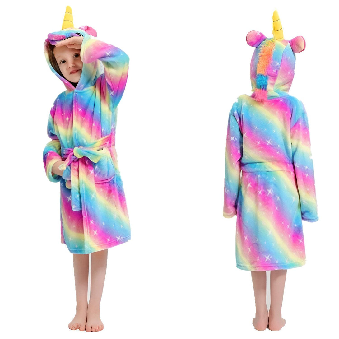 Une jeune fille porte une robe de chambre licorne arc-en-ciel avec une capuche. La robe de chambre est en peluche et a des étoiles blanches. La capuche a une corne de licorne et des oreilles de licorne. La jeune fille est debout et regarde vers la caméra.