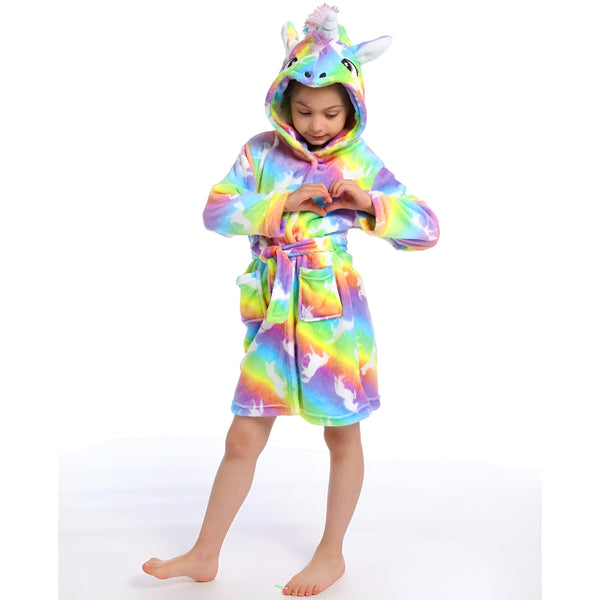 Une jeune fille porte une robe de chambre licorne arc-en-ciel avec une capuche. La robe de chambre est en peluche et a une ceinture. La fille a les cheveux longs et blonds et elle sourit.