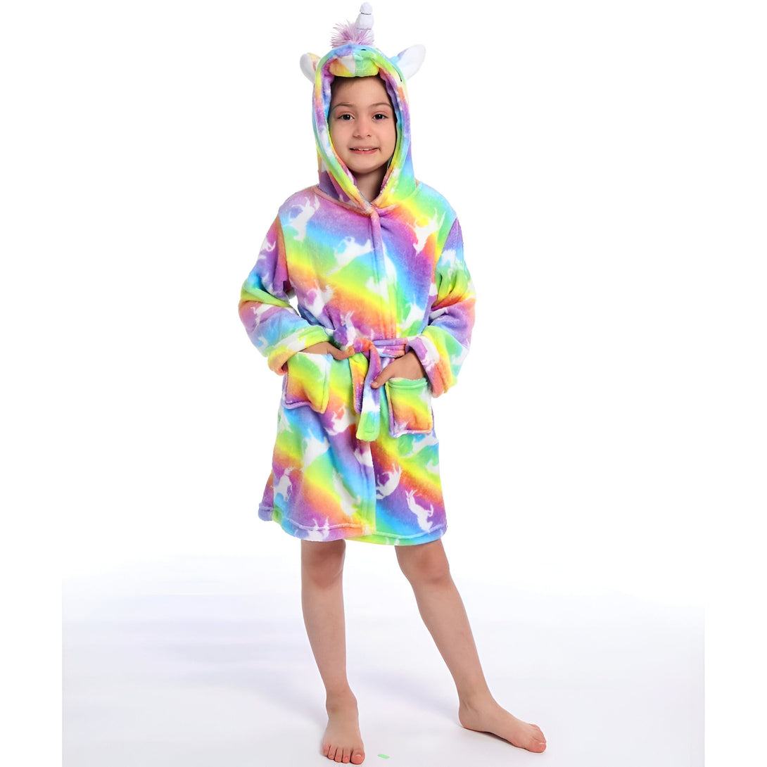 Une jeune fille porte une robe de chambre licorne arc-en-ciel avec une capuche. La robe de chambre est en peluche et a une ceinture. La capuche a une corne de licorne et des oreilles de licorne. La jeune fille a les cheveux longs et blonds et elle sourit.