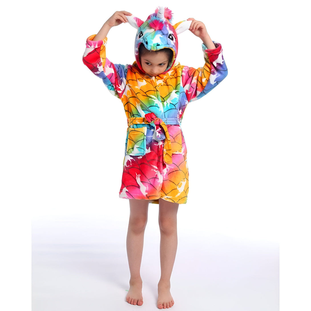 Une jeune fille porte une robe de chambre licorne arc-en-ciel. La robe de chambre a une capuche avec une tête de licorne et des oreilles. La robe de chambre est attachée avec une ceinture. La jeune fille est pieds nus.