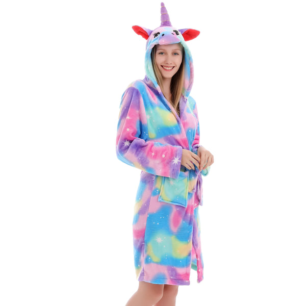 Une jeune femme porte un peignoir licorne. Le peignoir est rose, violet et bleu avec une capuche en forme de tête de licorne. La femme est souriante et a les cheveux longs.