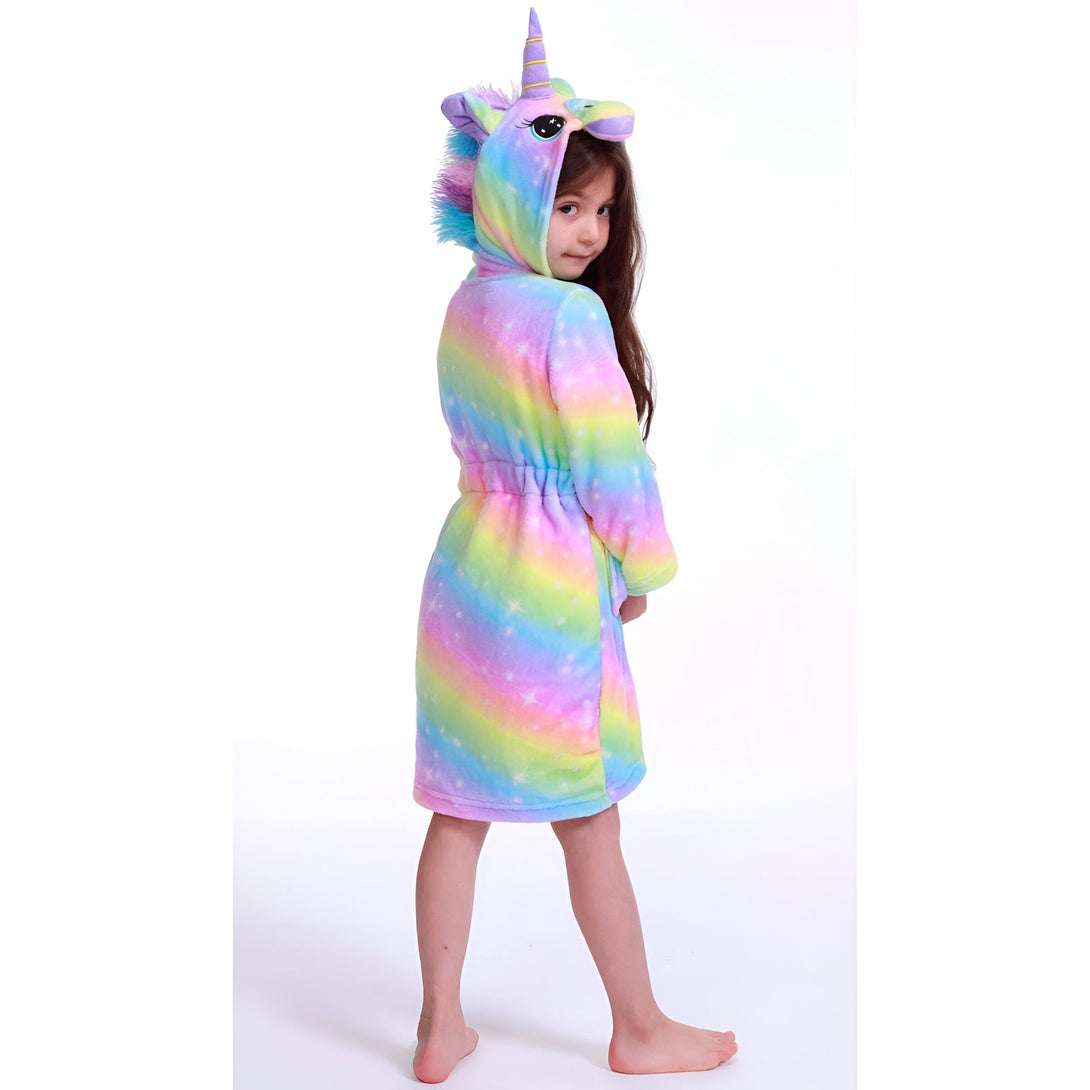 Une jeune fille porte une robe de chambre licorne arc-en-ciel avec une capuche. La robe de chambre est en peluche et a des étoiles et des lunes imprimées. La capuche a une corne de licorne et des oreilles.