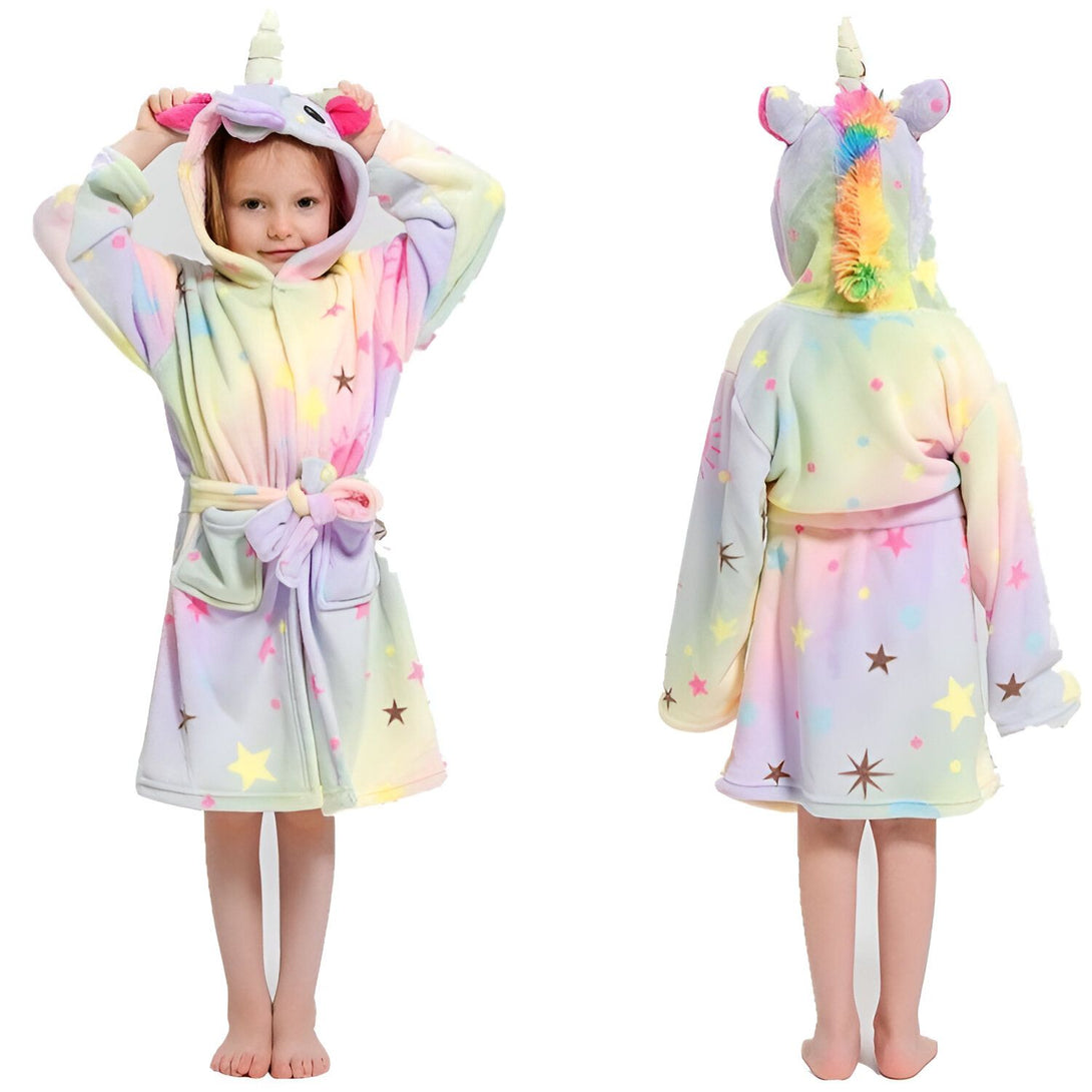 Une petite fille porte une robe de chambre licorne. La robe de chambre est rose, violette et bleue avec des étoiles et une capuche avec une corne de licorne. La petite fille a les cheveux blonds et elle est souriante.