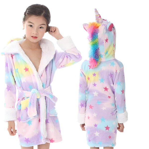 Une jeune fille porte une robe de chambre licorne. La robe de chambre est rose et violette avec des étoiles et des arcs-en-ciel. La capuche de la robe de chambre a la forme d'une tête de licorne avec une corne et des oreilles.