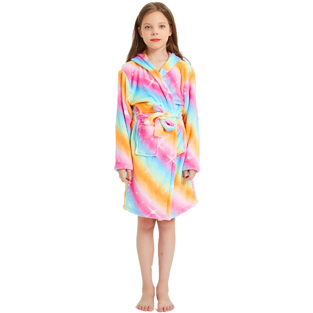 Une jeune fille porte une robe de chambre arc-en-ciel à capuche. La robe de chambre est en peluche et a une ceinture. La fille a les cheveux bruns et les yeux marrons. Elle est pieds nus et se tient debout sur un sol blanc.