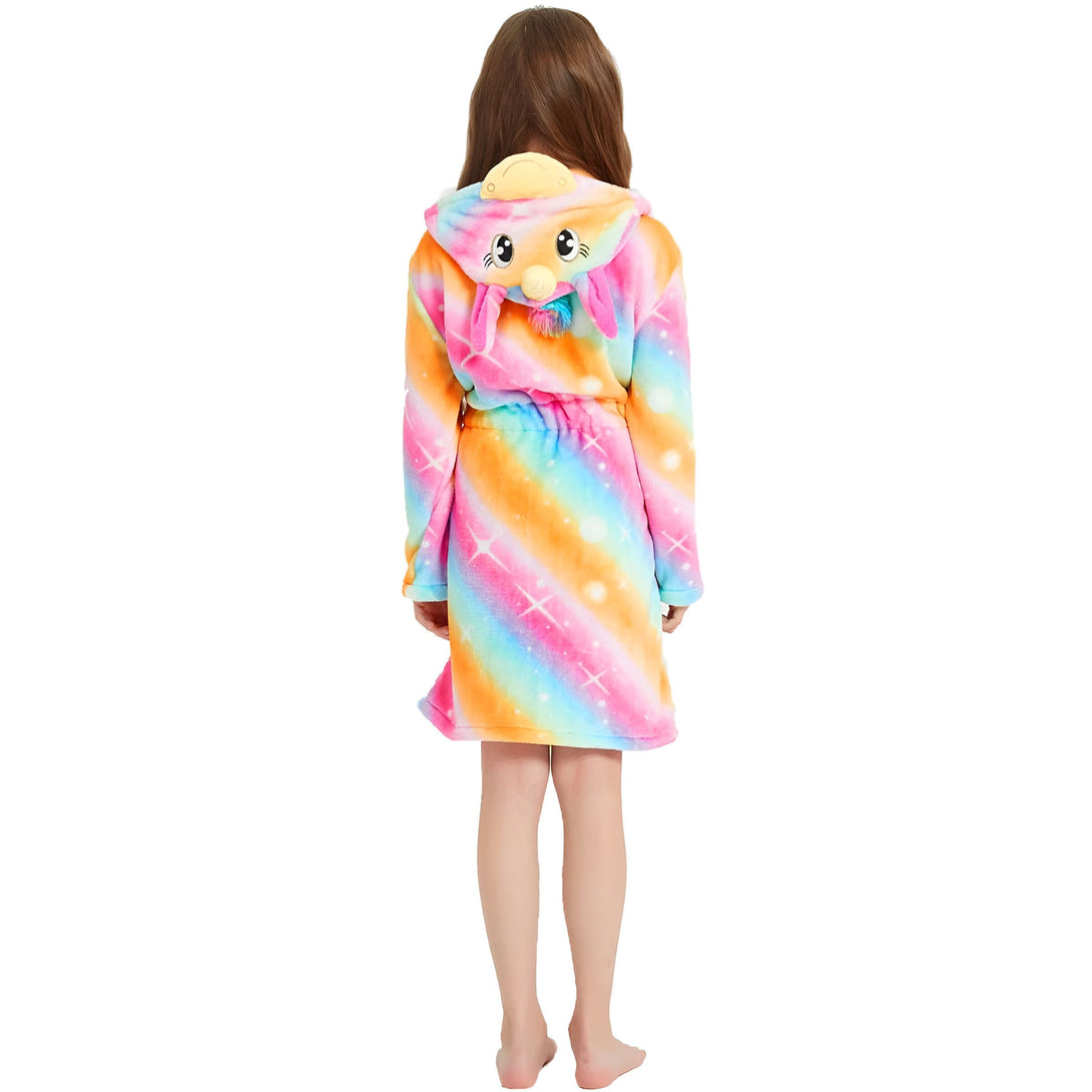 Une jeune fille porte une robe de chambre licorne arc-en-ciel avec une capuche. La robe de chambre est en peluche et a des étoiles et des lunes imprimées. La capuche a une corne de licorne et des oreilles de lapin.