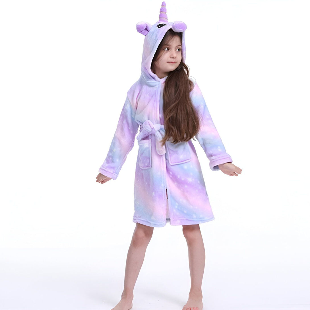 Une jeune fille porte une robe de chambre licorne violette et blanche. La robe de chambre a une capuche avec une corne de licorne et des oreilles. La robe de chambre est également dotée d'une ceinture.