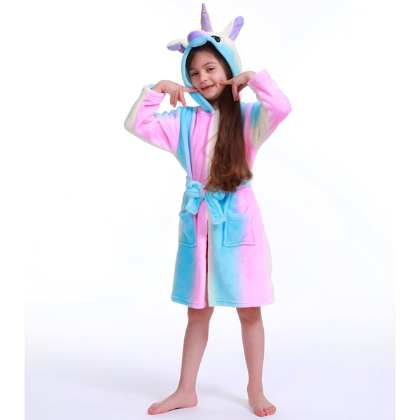 Une jeune fille porte une combinaison pyjama licorne avec une capuche. La combinaison est rose, bleue et violette. La capuche est en forme de tête de licorne avec une corne et des oreilles. La combinaison est attachée avec une ceinture. La jeune fille est pieds nus et a les cheveux longs.