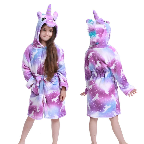 Une jeune fille porte une combinaison pyjama licorne violette avec une capuche. La combinaison est à manches longues et possède une ceinture à la taille. La capuche est ornée d'une crinière et d'une corne de licorne.