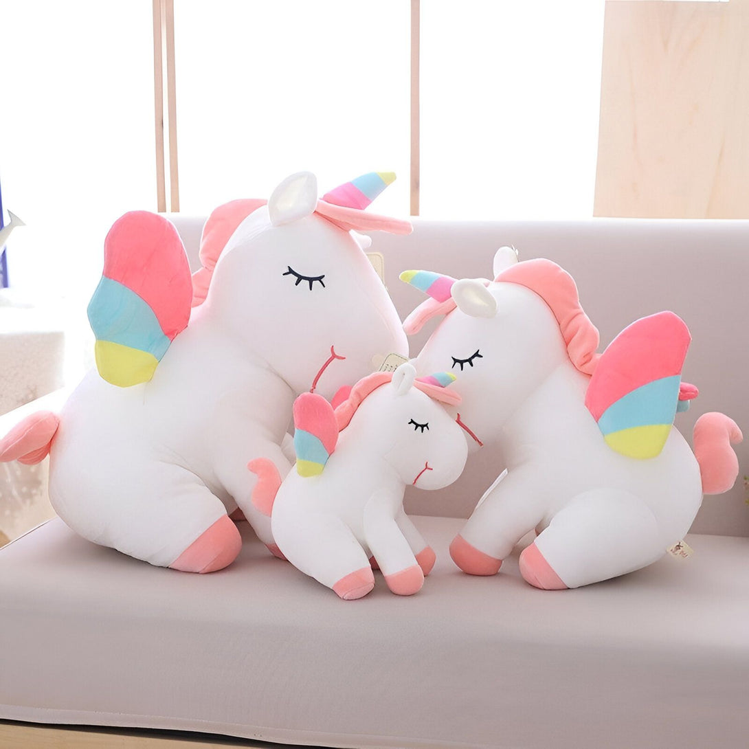 Une peluche licorne blanche avec des ailes arc-en-ciel, une crinière et une queue roses. Elle est allongée sur un canapé avec ses deux petits.