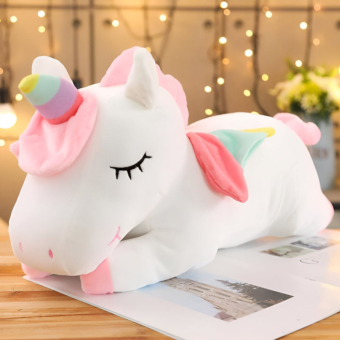 Une peluche licorne blanche et rose allongée sur une table. La peluche a une crinière et une queue arc-en-ciel. Elle est très mignonne et a l'air très douce.