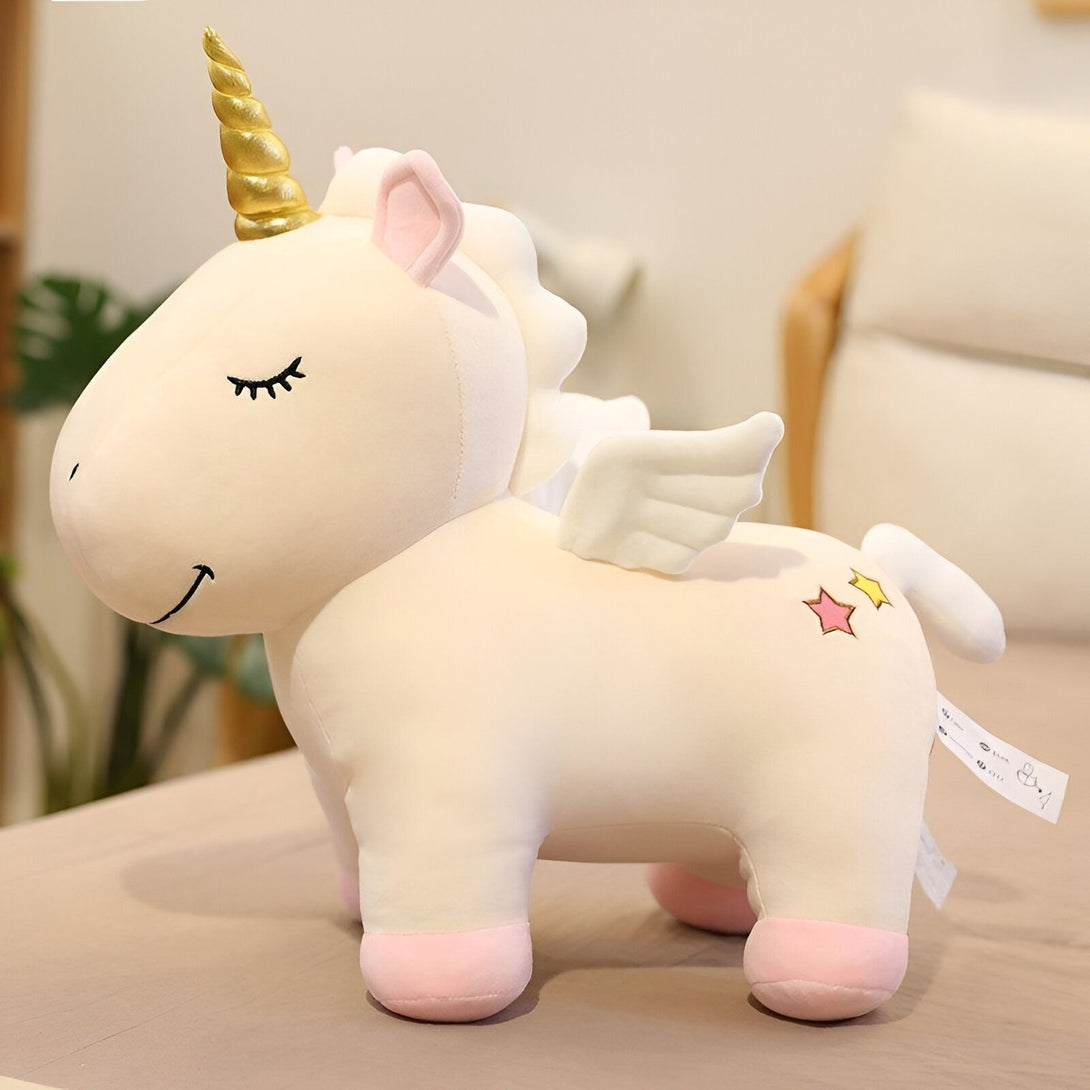 Une peluche licorne blanche avec une crinière et une queue roses, des étoiles sur la croupe, des ailes et une corne dorées.