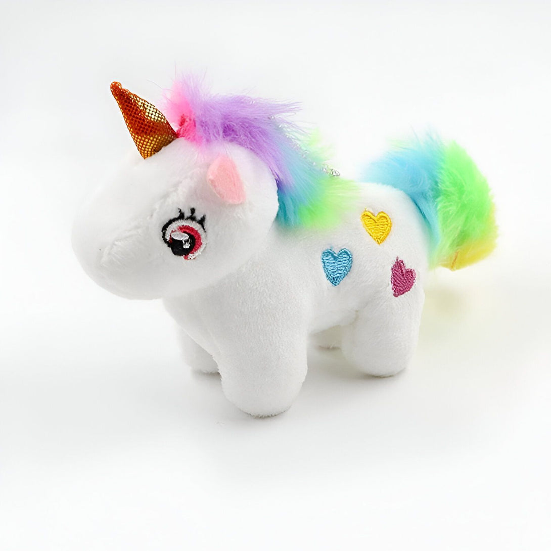 Une peluche de licorne blanche avec une crinière et une queue arc-en-ciel. Elle a une corne dorée et des yeux roses. Il y a des coeurs multicolores sur son corps.