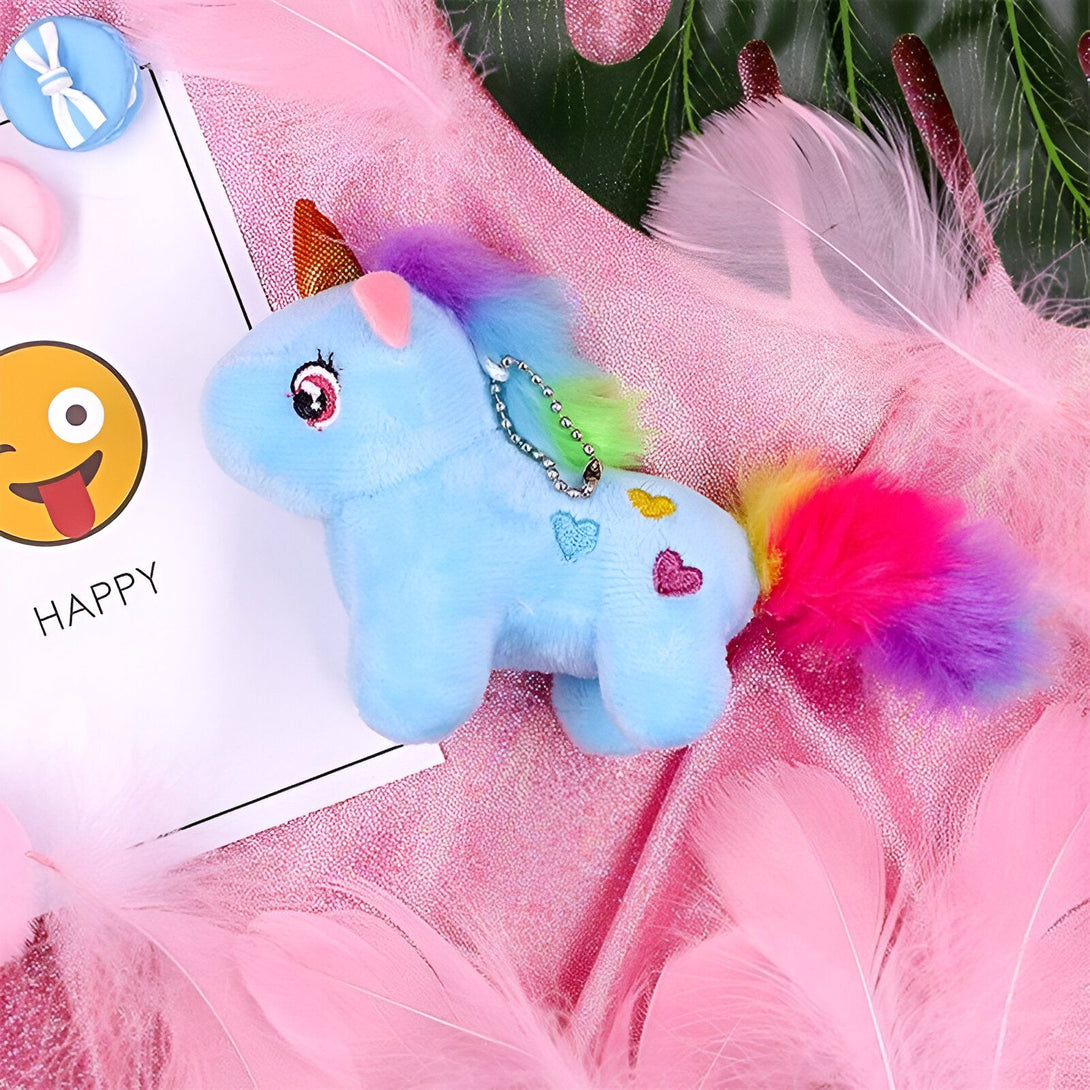 Une peluche licorne bleue avec une crinière et une queue arc-en-ciel. Elle a une corne dorée et des yeux roses. Elle est posée sur un fond rose avec des plumes blanches et un visage souriant jaune.