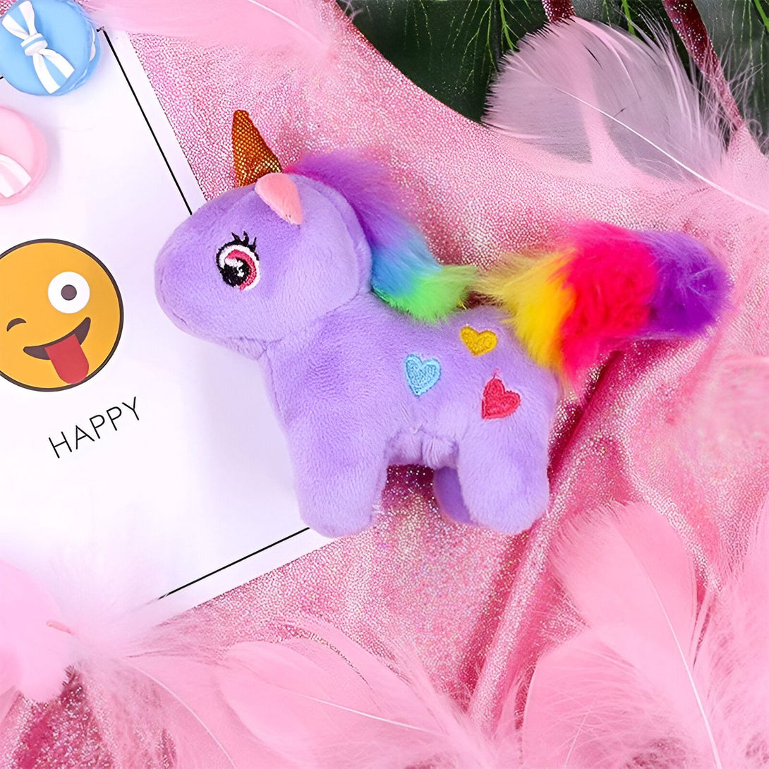 Une peluche licorne violette avec une crinière et une queue arc-en-ciel. La peluche est posée sur un fond de plumes roses.