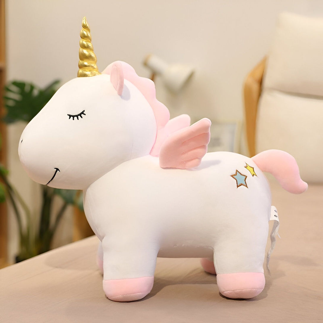 Une peluche licorne blanche et rose avec une corne dorée et des ailes roses. La peluche est posée sur une table.