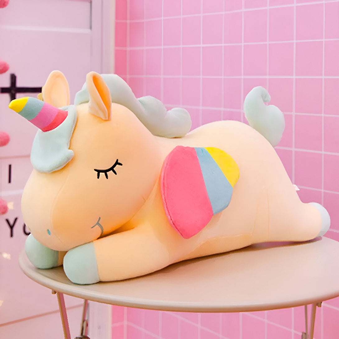 Une peluche licorne jaune allongée sur une table avec les yeux fermés. La peluche a une crinière et une queue arc-en-ciel, ainsi que des ailes roses et bleues.