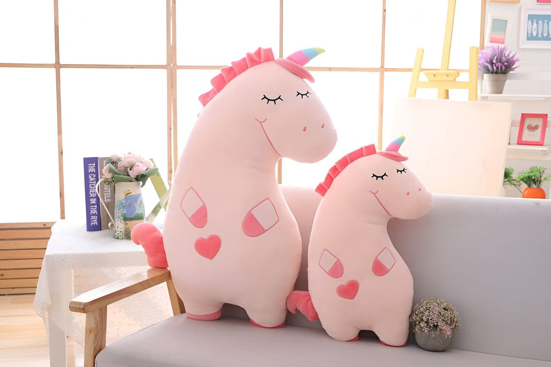 Deux peluches licornes roses, une grande et une petite, sont assises sur un canapé gris clair devant une fenêtre ensoleillée. Les licornes ont des crinières roses, des cornes multicolores et des cœurs roses sur leur corps., 45501421551871 | Licorne Shop
