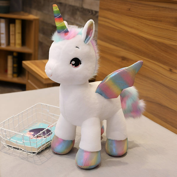 Une peluche licorne blanche avec une crinière et une queue arc-en-ciel. Elle a des ailes irisées et des sabots argentés.