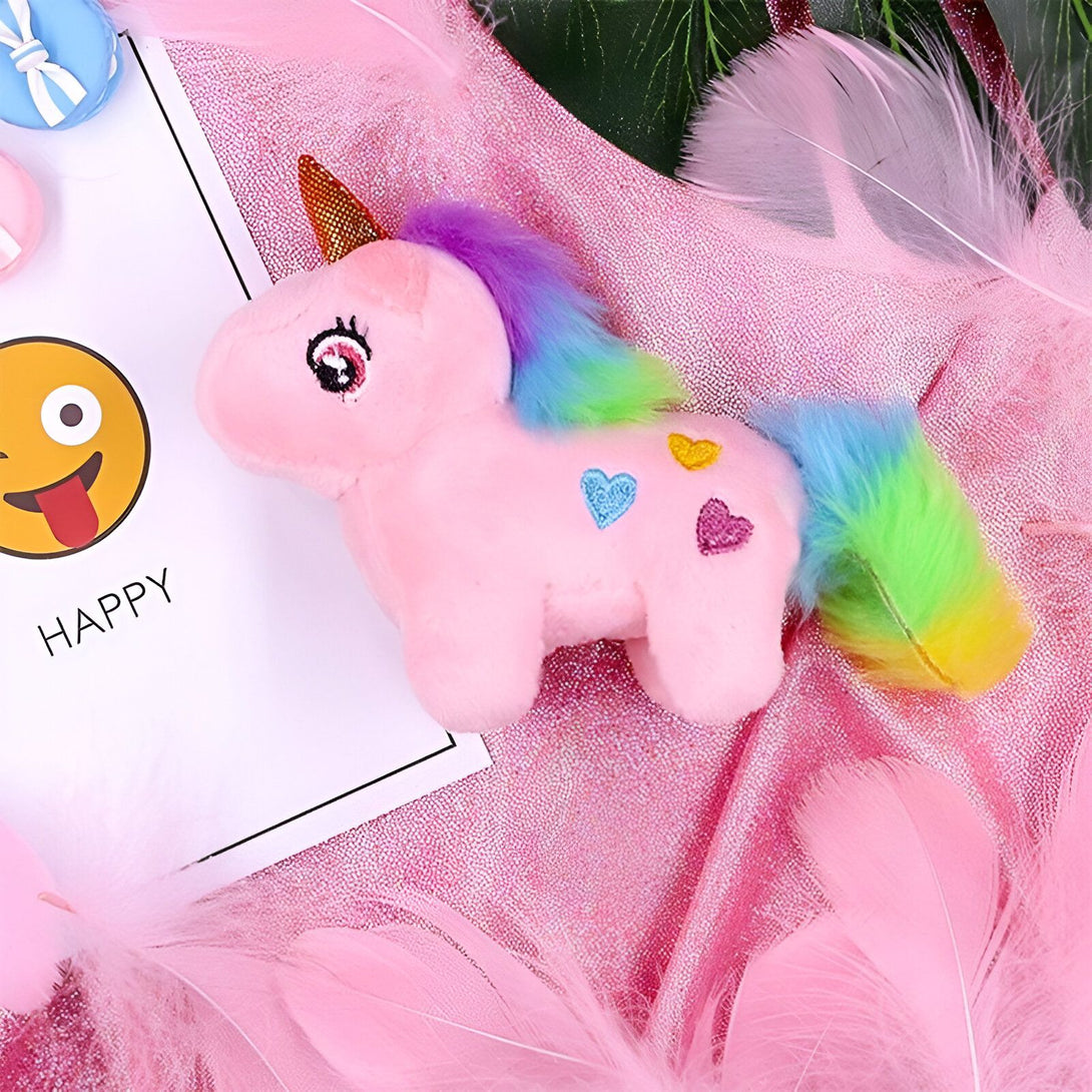 Une peluche licorne rose avec une crinière arc-en-ciel et des cœurs sur le corps. Elle est posée sur un fond rose pailleté.