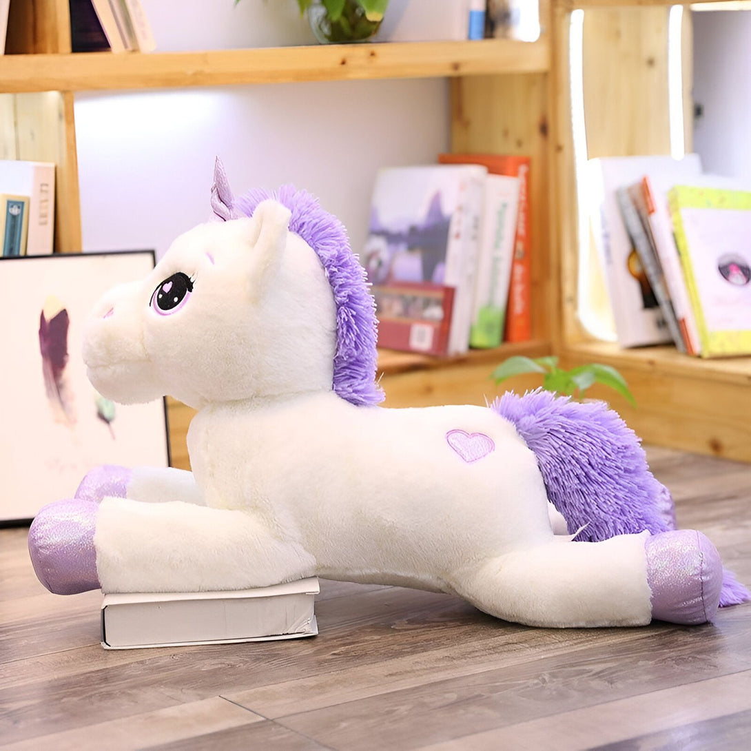 Une peluche licorne blanche et violette allongée sur le sol devant une bibliothèque.