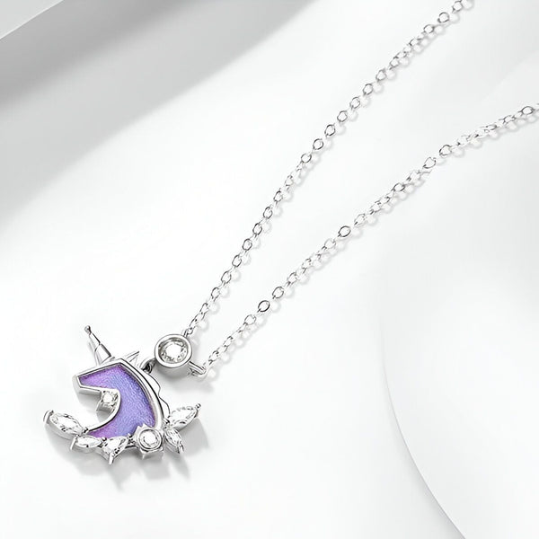 Un collier avec un pendentif en forme de licorne. La licorne est violette avec une crinière et une queue roses. Le pendentif est entouré de feuilles et de fleurs. La chaîne est en argent.