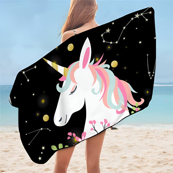 Une femme sur la plage enveloppée dans une serviette noire imprimée d'une licorne blanche avec une crinière rose et bleue, des étoiles et des constellations dorées., 45501225763071 | Licorne Shop
