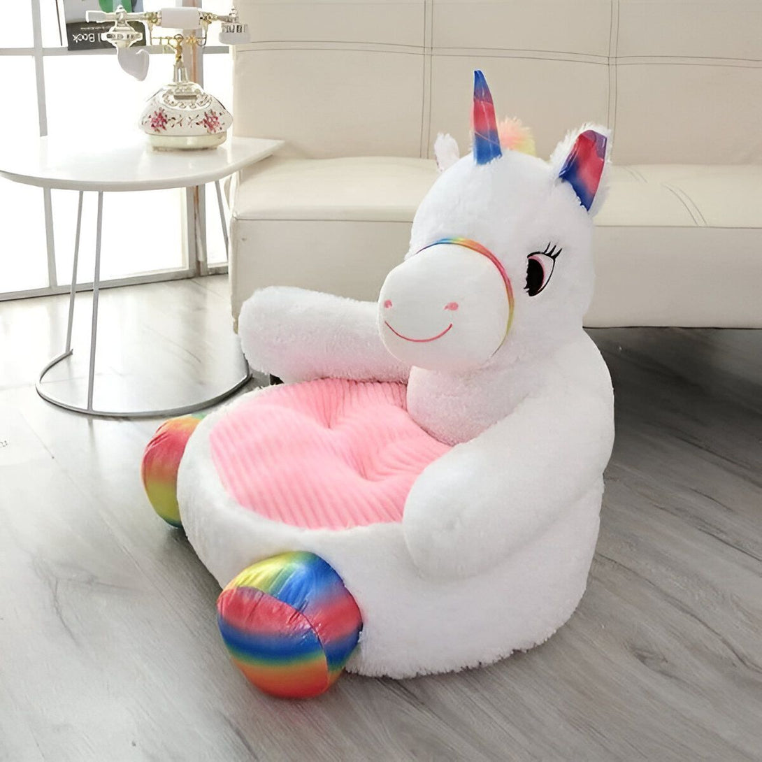 Une peluche licorne blanche avec une crinière et une queue arc-en-ciel. Elle est assise sur un siège rembourré rose avec des accoudoirs et un dossier. La peluche est souriante et a les yeux bleus. Elle est destinée aux enfants.