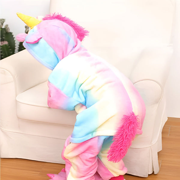 Enfant portant un pyjama licorne multicolore avec capuche, corne jaune et crinière rose. Le costume a une queue rose touffue. L'enfant est debout devant un fauteuil de couleur crème., 45502353080575,45502353113343,45502353146111,45502353178879,45502353211647,45502353244415,45502353277183,45502353309951 | Licorne Shop