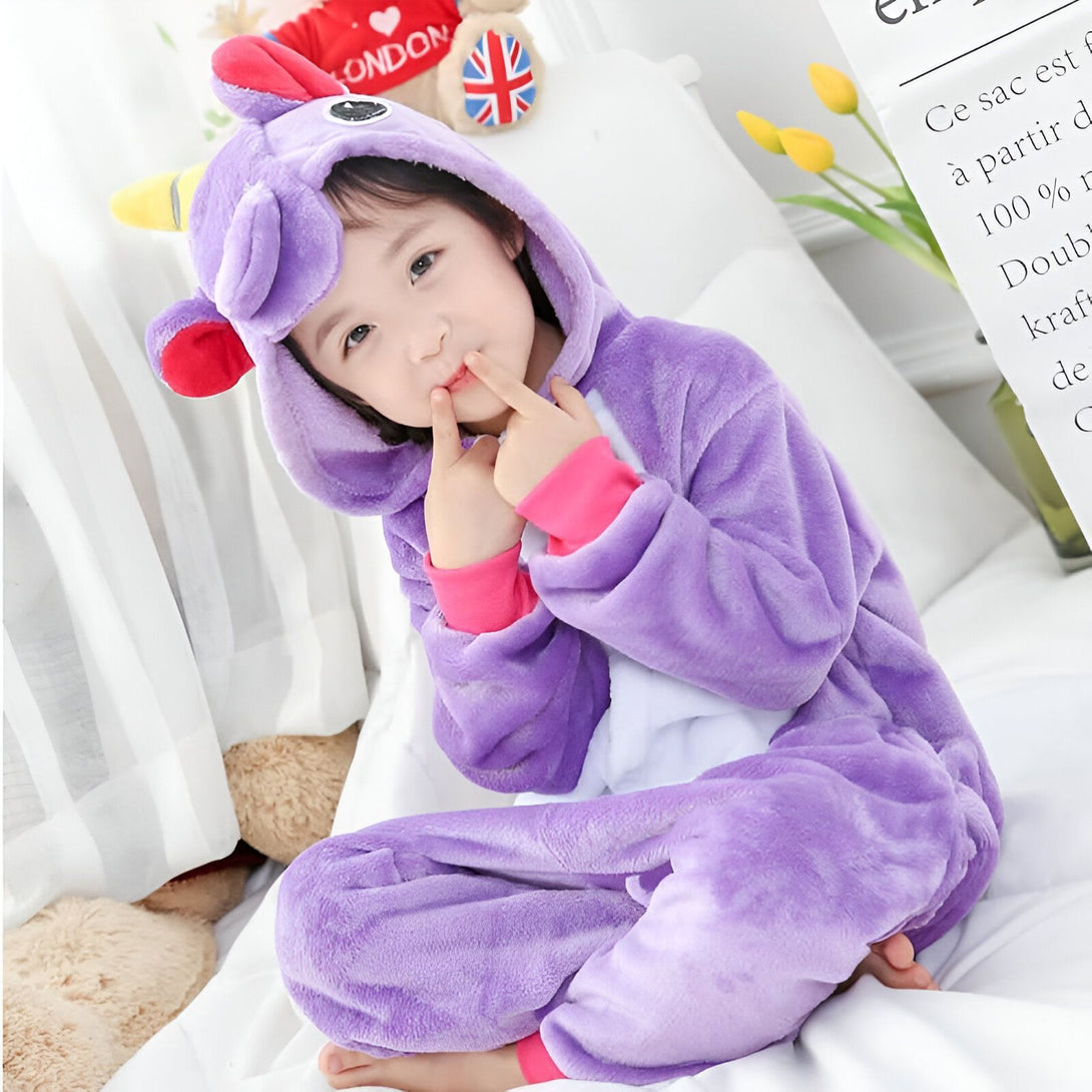 Une petite fille assise sur un lit, portant un pyjama violet en forme de licorne avec une capuche., 45502354751743,45502354784511,45502354817279,45502354850047,45502354882815,45502354915583,45502354948351,45502354981119 | Licorne Shop