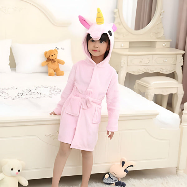 Une petite fille en peignoir rose licorne dans sa chambre. Un ourson en peluche et d'autres jouets sont sur le lit. Une coiffeuse blanche est visible en arrière-plan., 45502030971135,45502031003903,45502031036671,45502031069439,45502031102207,45502031134975,45502031167743,45502031200511 | Licorne Shop