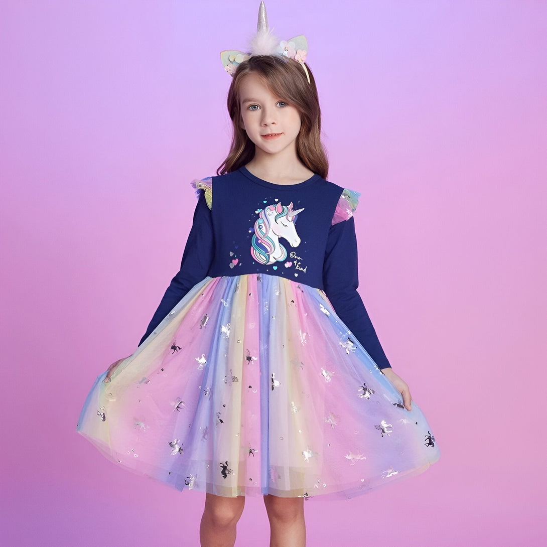 Une jeune fille porte une robe à manches longues bleue avec un motif de licorne. La robe est ornée de tulle arc-en-ciel et de paillettes. Elle porte également un serre-tête avec des oreilles de licorne.