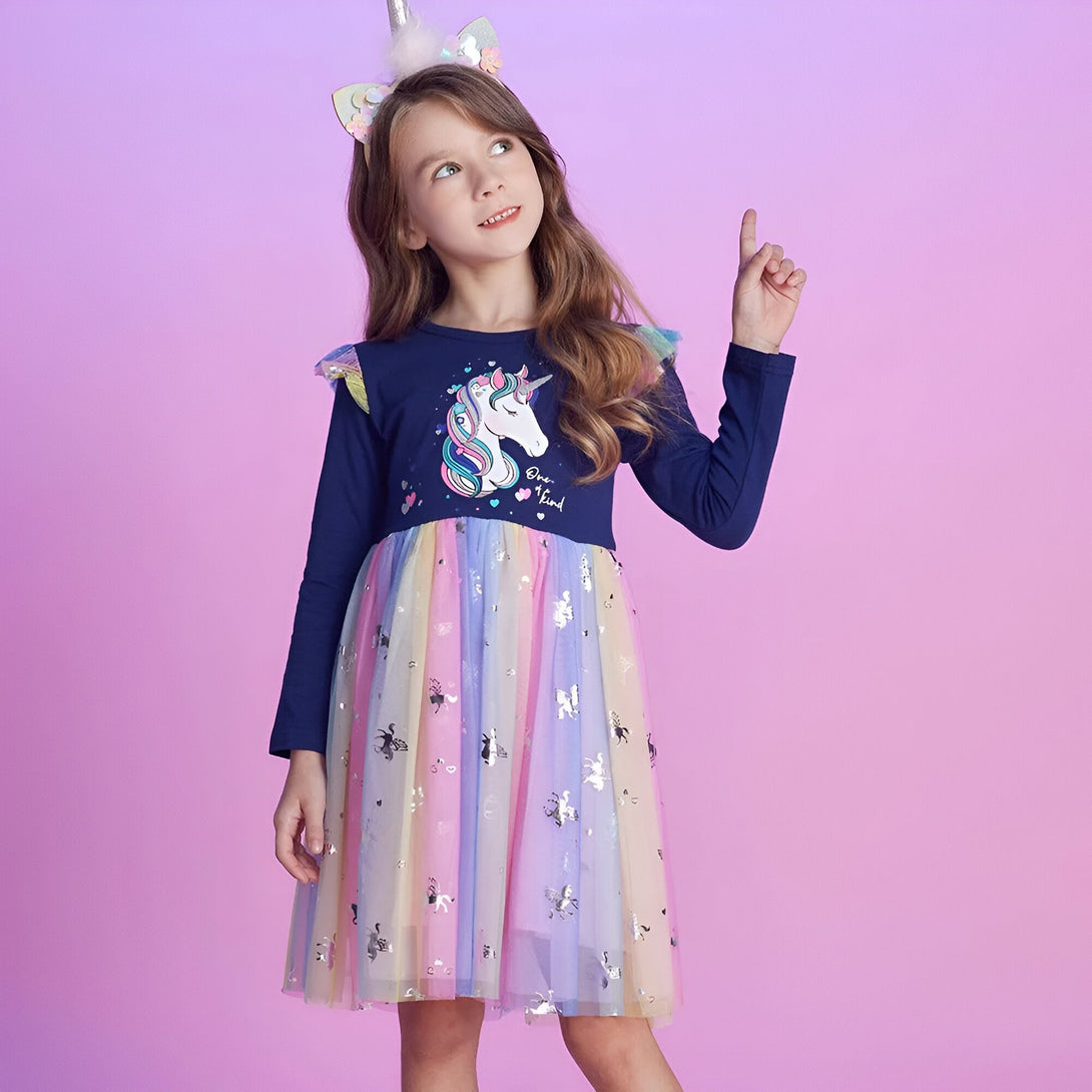 Une fille porte une robe à manches longues avec une licorne imprimée sur le devant. La robe est bleue et rose avec un tutu arc-en-ciel. La fille a un serre-tête avec des oreilles de licorne et pointe du doigt.