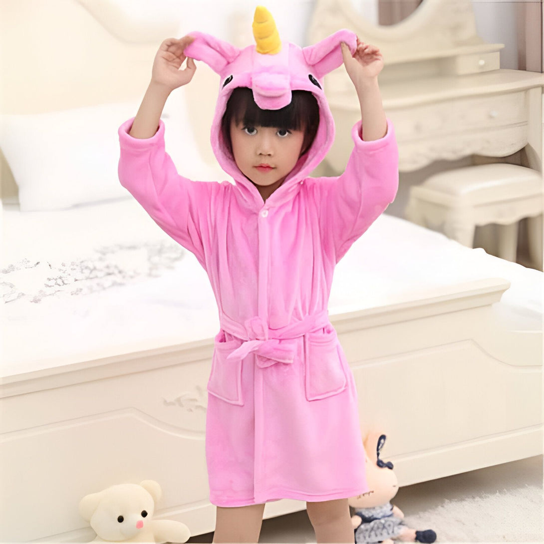 Enfant portant un peignoir rose à capuche en forme de licorne, debout devant un lit et une commode blanche. Deux jouets en peluche, un ours et une poupée, se trouvent à ses pieds., 45502348034303,45502348067071,45502348099839,45502348132607,45502348165375,45502348198143,45502348230911,45502348263679 | Licorne Shop
