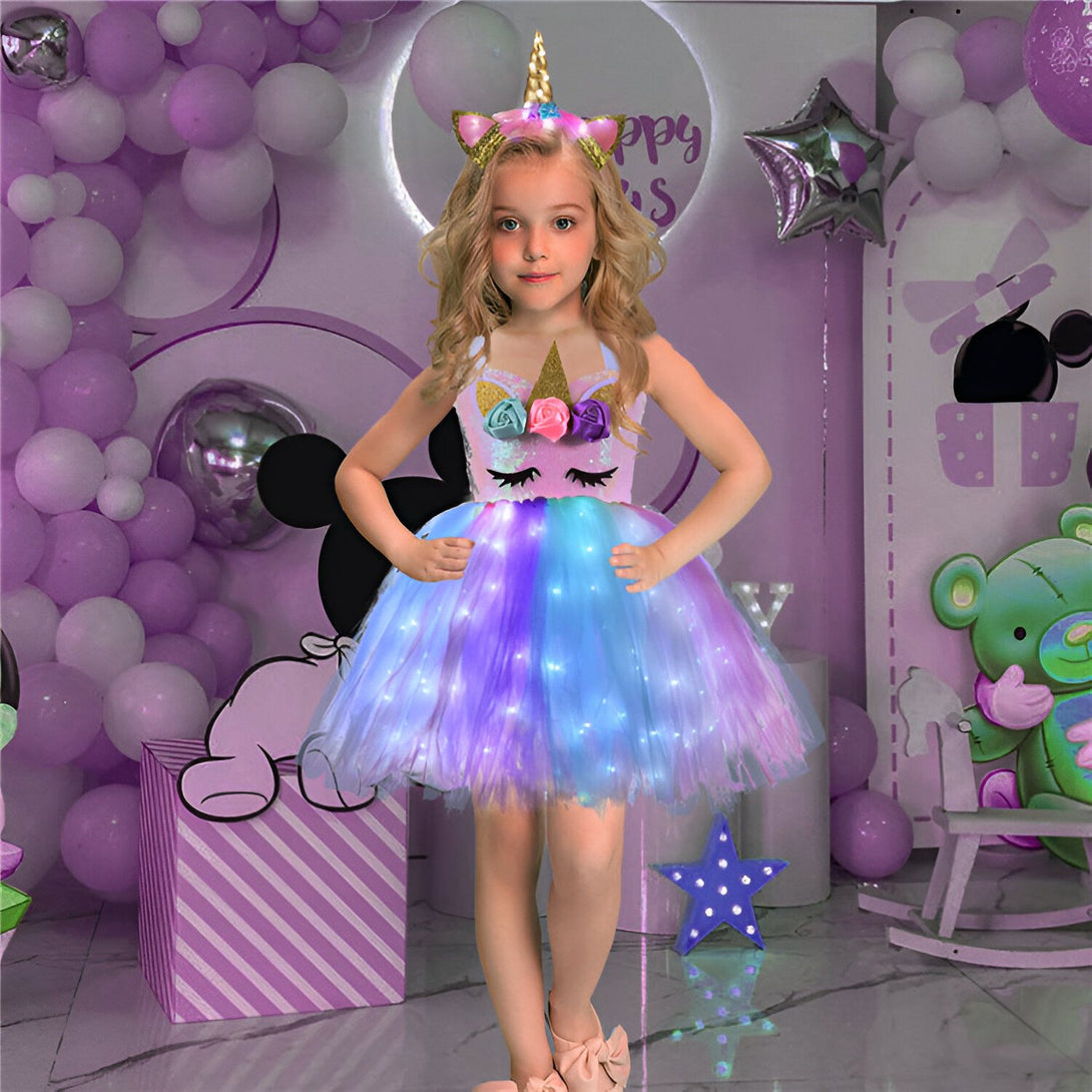 Une petite fille porte une robe de licorne avec des lumières LED. La robe est rose et violette et a une jupe en tulle. La fille a un serre-tête avec une licorne et des fleurs. Elle se tient dans un décor de fête avec des ballons et des peluches.