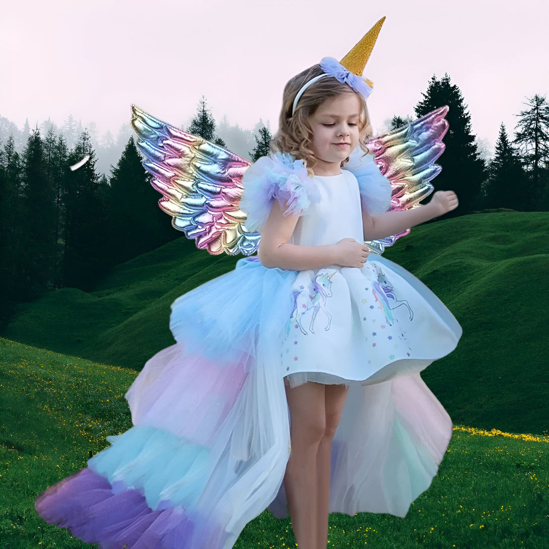 Une petite fille porte une robe de licorne blanche et arc-en-ciel. Elle a des ailes de licorne en plastique et une couronne de licorne en plastique sur la tête. Elle se tient dans un champ d'herbe verte avec des arbres en arrière-plan.