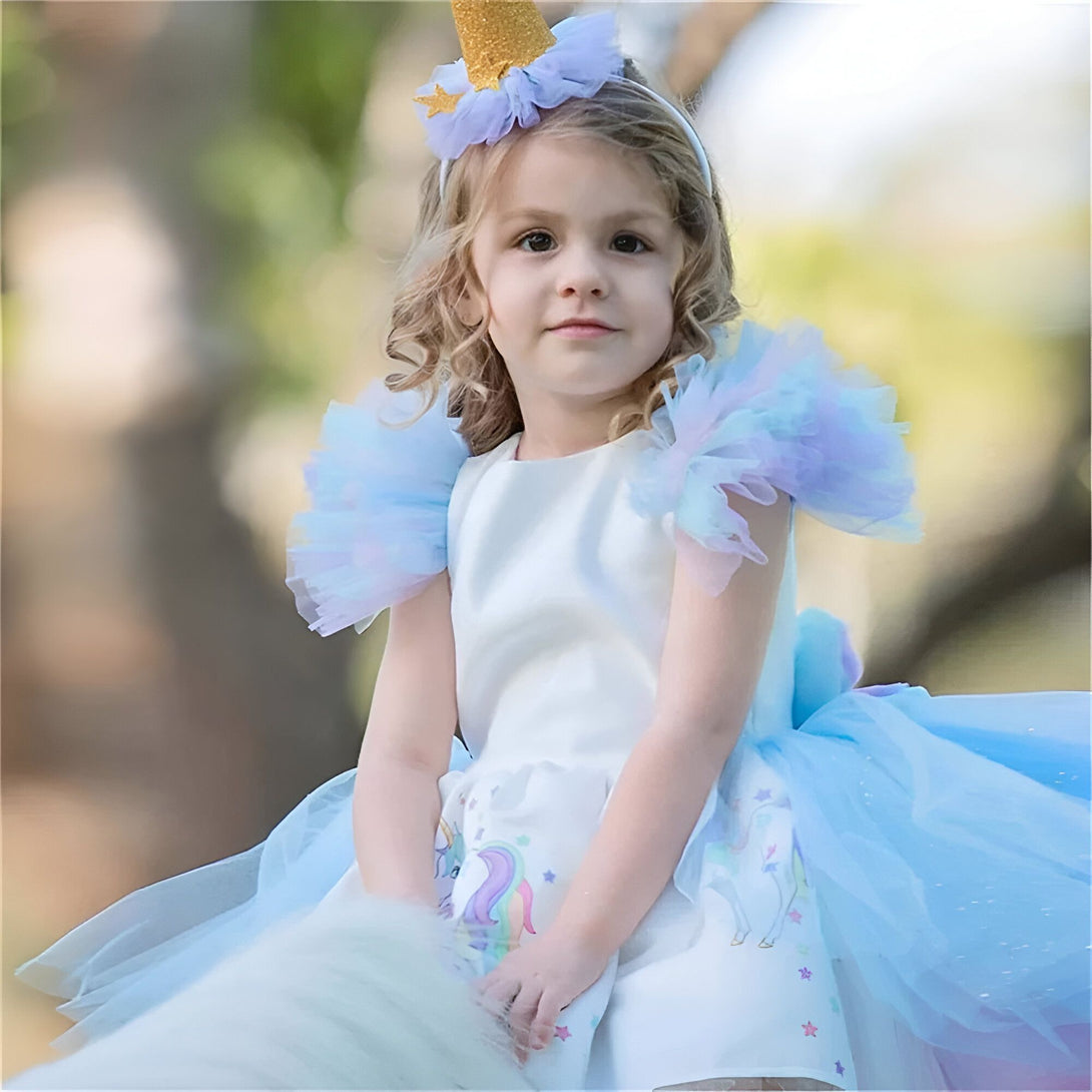 Une petite fille porte une robe de licorne blanche et bleue. Elle a une couronne de fleurs sur la tête et un serre-tête avec des oreilles de licorne. Elle est assise sur un poney et sourit à l'appareil photo.