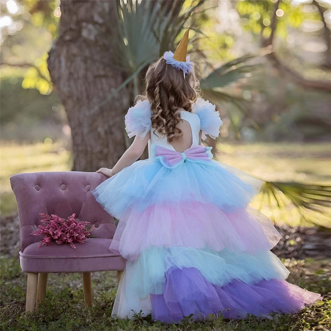 Une petite fille porte une robe de licorne blanche, rose et bleue. Elle a une couronne de licorne sur la tête et un nœud dans le dos. Elle est assise sur une chaise dans un jardin.