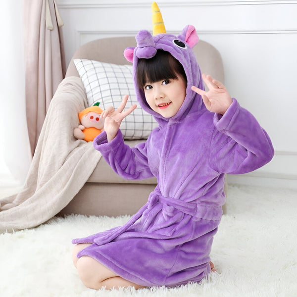 Une petite fille souriante à genoux sur un tapis blanc, portant un peignoir violet à capuche avec une corne de licorne et des oreilles roses. Elle fait un signe de paix avec ses mains. Derrière elle, on aperçoit un canapé, un coussin et une peluche orange., 45502350196991,45502350229759,45502350262527,45502350295295,45502350328063,45502350360831,45502350393599,45502350426367 | Licorne Shop