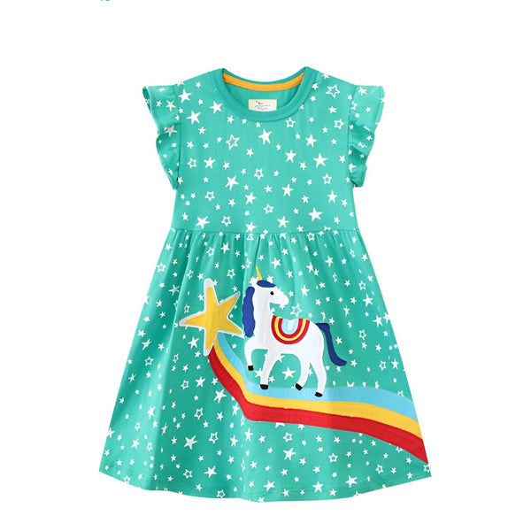 Une petite fille porte une robe verte à manches courtes. La robe est imprimée d'étoiles blanches et d'une licorne. La licorne est blanche avec une crinière et une queue arc-en-ciel. La robe est évasée et s'arrête au-dessus du genou.