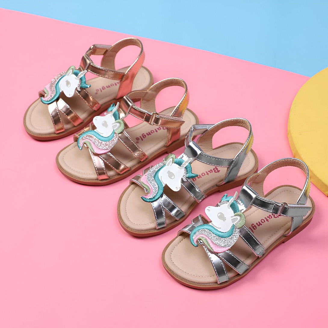 Une paire de sandales à scratchs pour enfant avec une licorne sur le dessus. Les sandales sont disponibles en trois couleurs : rose, argent et doré.