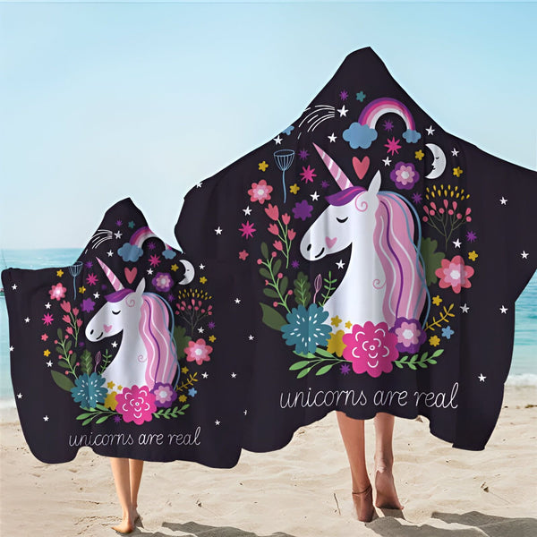 Deux personnes sur une plage portant des serviettes à capuche à motif de licorne. Les serviettes sont noires et décorées de licornes blanches avec des crinières roses et de fleurs colorées. Le texte "unicorns are real" est visible., 45501225238783 | Licorne Shop